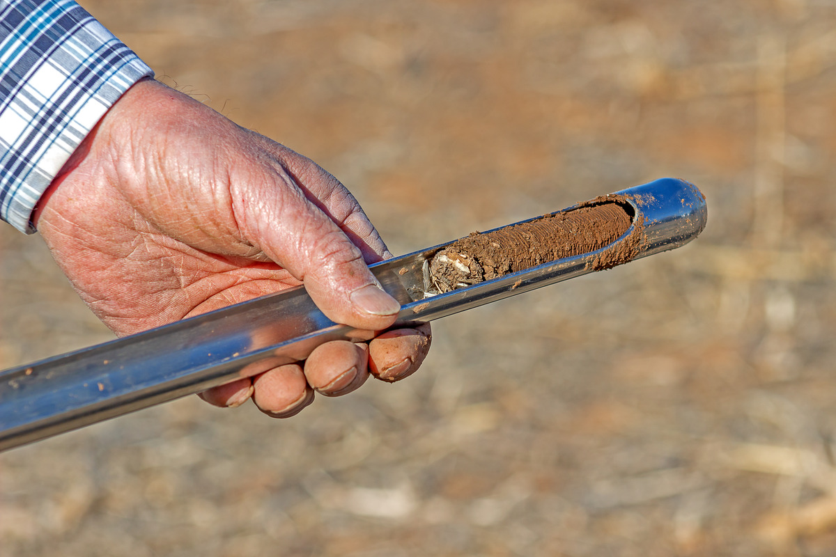 A man wearing a blue and white checked shirt holding a metal tool used for soil sampling, with a cylinder of dry soil inside.