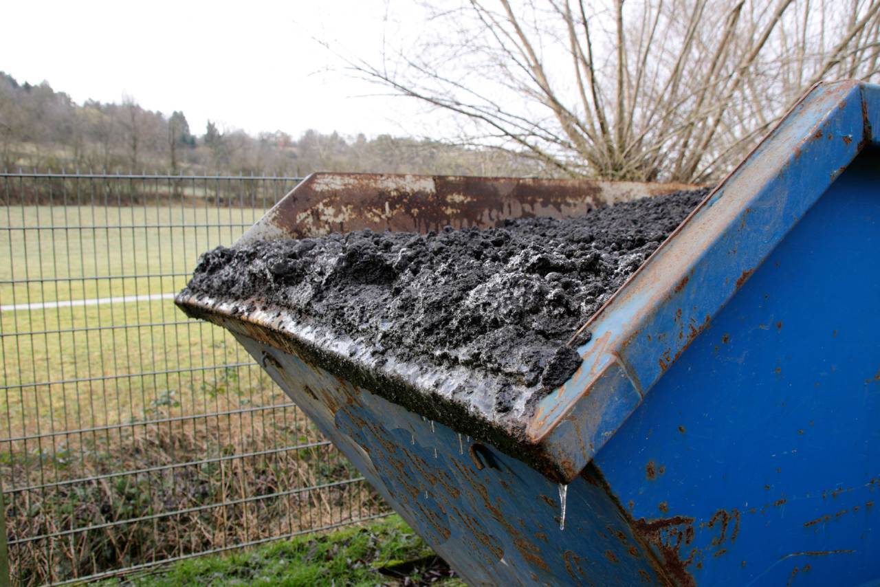 A blue container filled with wet, dark sludge placed near a wire fence. In the background, a grassy field and leafless trees.