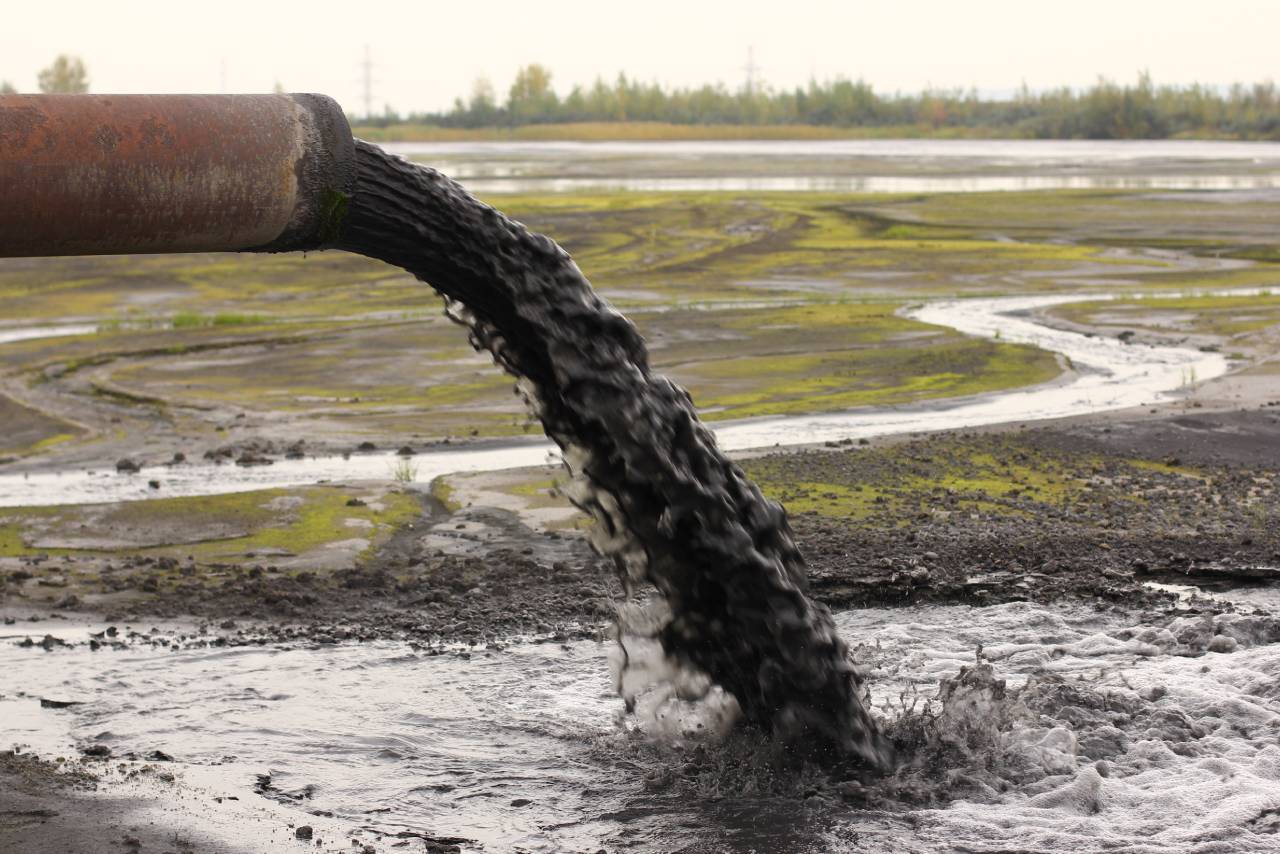 An industrial pipe discharges thick, dark liquid waste into a muddy landscape with green patches, with an overcast sky in the background.